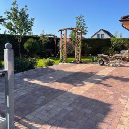 La-Bud usługi budowlane - Ułożona kostka brukowa w ogrodzie z drewnianymi pergolami, w tle zielony żywopłot i dom.