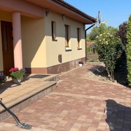 La-Bud usługi budowlane - Jasna elewacja domu jednorodzinnego z brązowymi akcentami, widoczne nowe brązowe płytki chodnikowe, kwiaty w donicach przy wejściu, w tle osoba pracująca w ogrodzie.