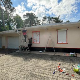 La-Bud usługi budowlane - Dwóch mężczyzn maluje elewację domu jednorodzinnego z dwoma garażami, okna zabezpieczone folią, narzędzia malarskie rozłożone na brukowanym podjeździe.
