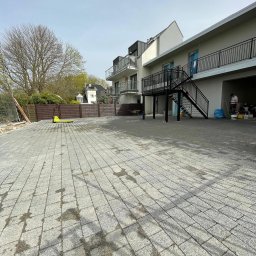 La-Bud usługi budowlane - Nowo ułożona szara kostka brukowa na posesji z dwupiętrowym budynkiem w tle, widoczny fragment ogrodzenia i pracownik w garażu.