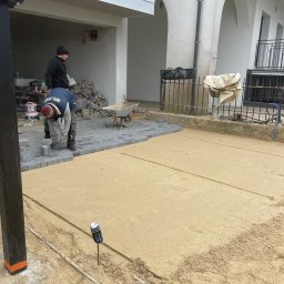 La-Bud usługi budowlane - Dwóch pracowników układających szarą kostkę brukową na podsypce piaskowej przed budynkiem z balkonami, widoczna taczka i zapas kostki.