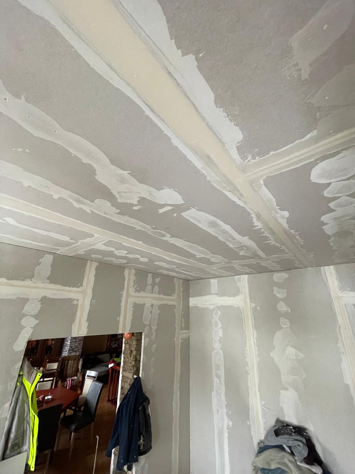 Wnętrze pokoju w trakcie remontu: ściany i sufit pokryte płytami karton gips, widoczne łączenia płyt wypełnione masą szpachlową, niezagruntowane powierzchnie, w tle widoczne meble w innym...