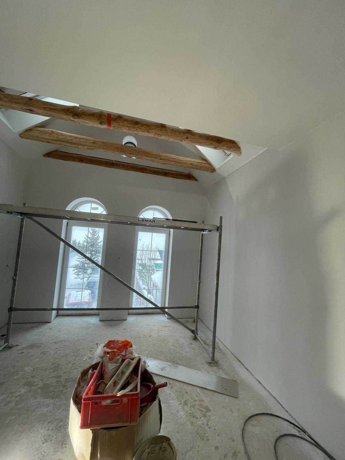Wnętrze w Pobierowie po szpachlowaniu ścian i sufitu, widoczne drewniane belki stropowe, rusztowanie przy oknach i narzędzia pracy.