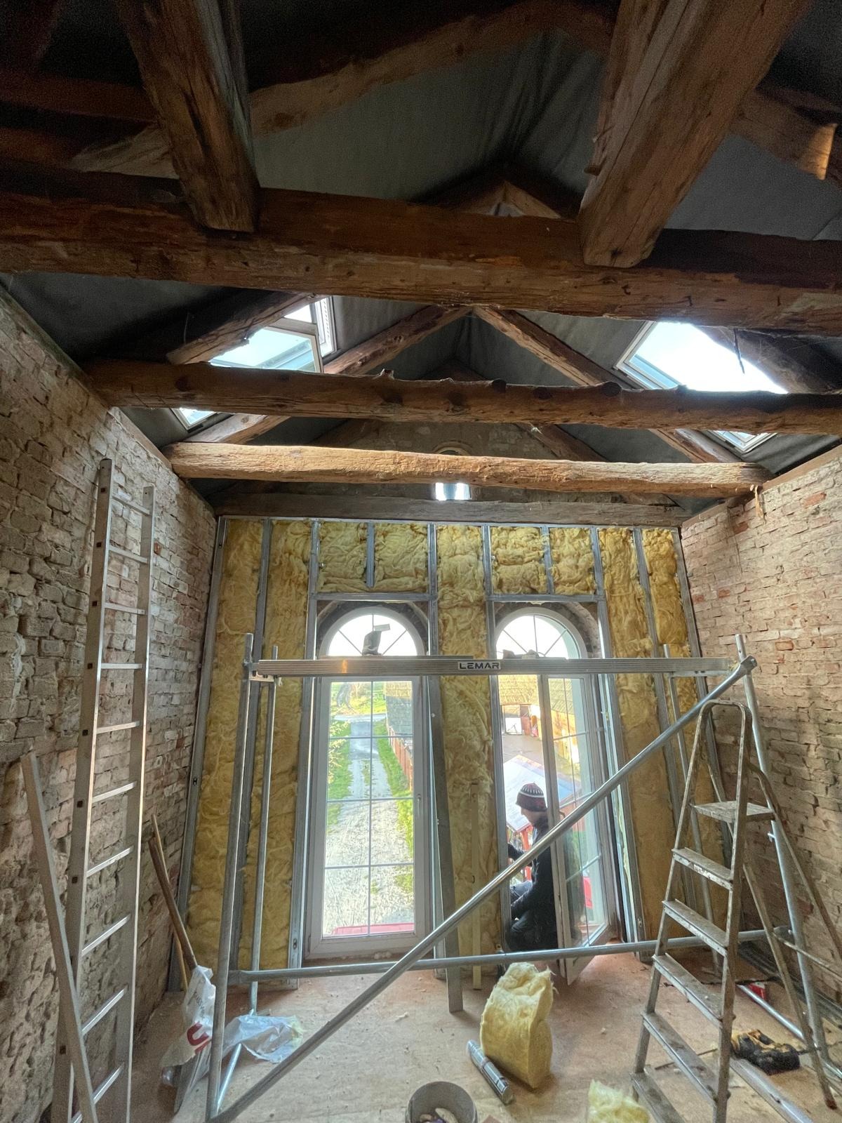 Wnętrze poddasza z odsłoniętą cegłą i drewnianymi belkami stropowymi, ocieplone wełną mineralną w metalowym stelażu, widoczne okna dachowe i drabiny.