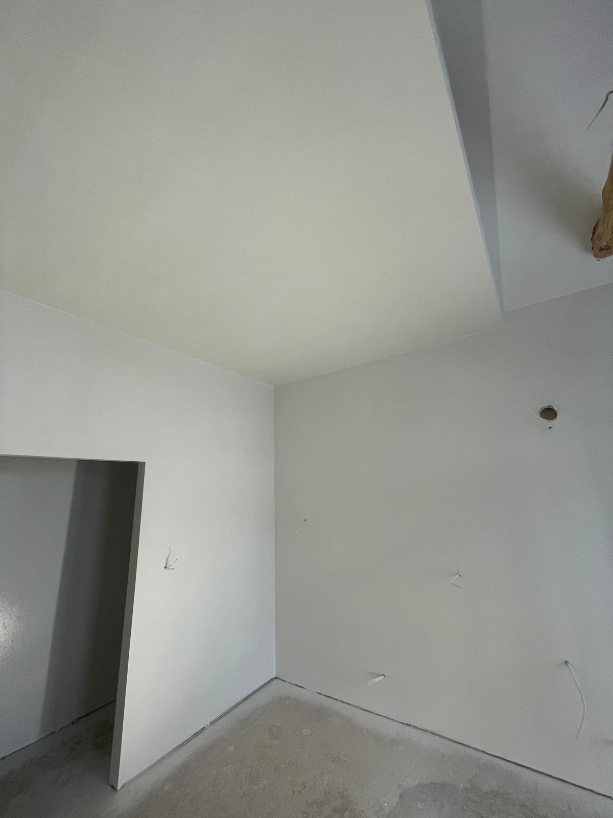 Wnętrze świeżo pomalowanego pokoju z widocznymi śladami po instalacjach elektrycznych i gładkim, białym wykończeniem ścian i sufitu.