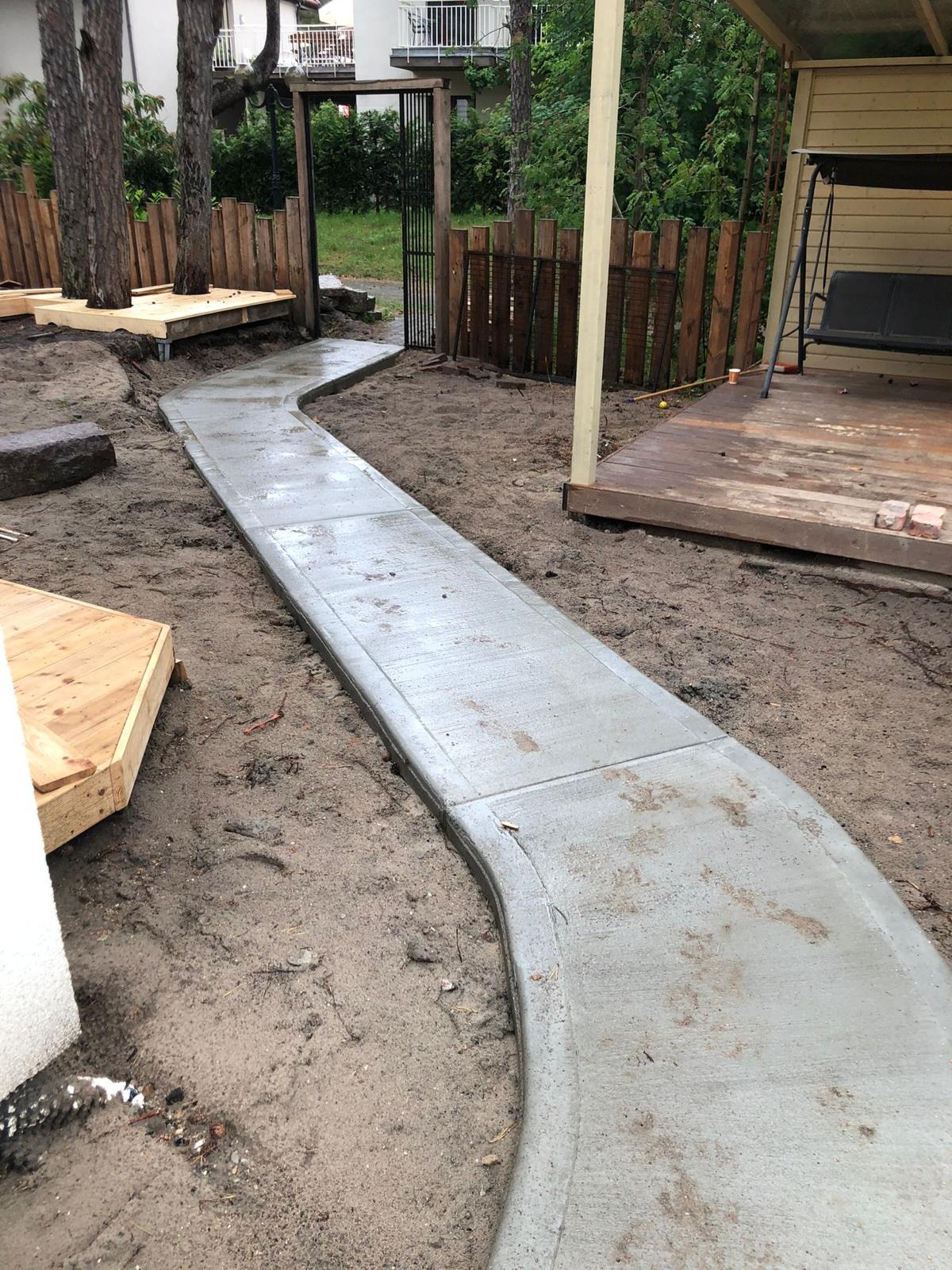 Świeżo wylana, betonowa ścieżka wijąca się przez ogród, prowadząca od drewnianego tarasu z huśtawką do metalowej bramki z drewnianym ogrodzeniem w tle. Wokół ścieżki widoczna świeżo rozkopana ziemia.