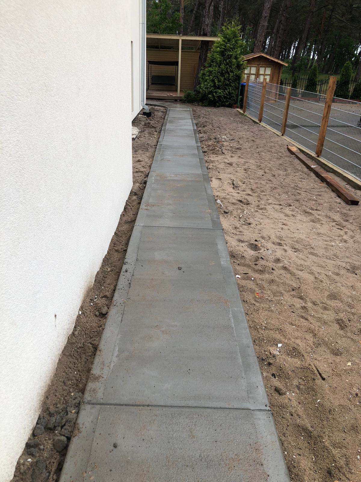 Świeżo ułożony, betonowy chodnik z szarych płyt prowadzący wzdłuż białej ściany budynku, z widocznym piaskiem i ziemią po bokach oraz fragmentem ogrodu z drzewami w tle.