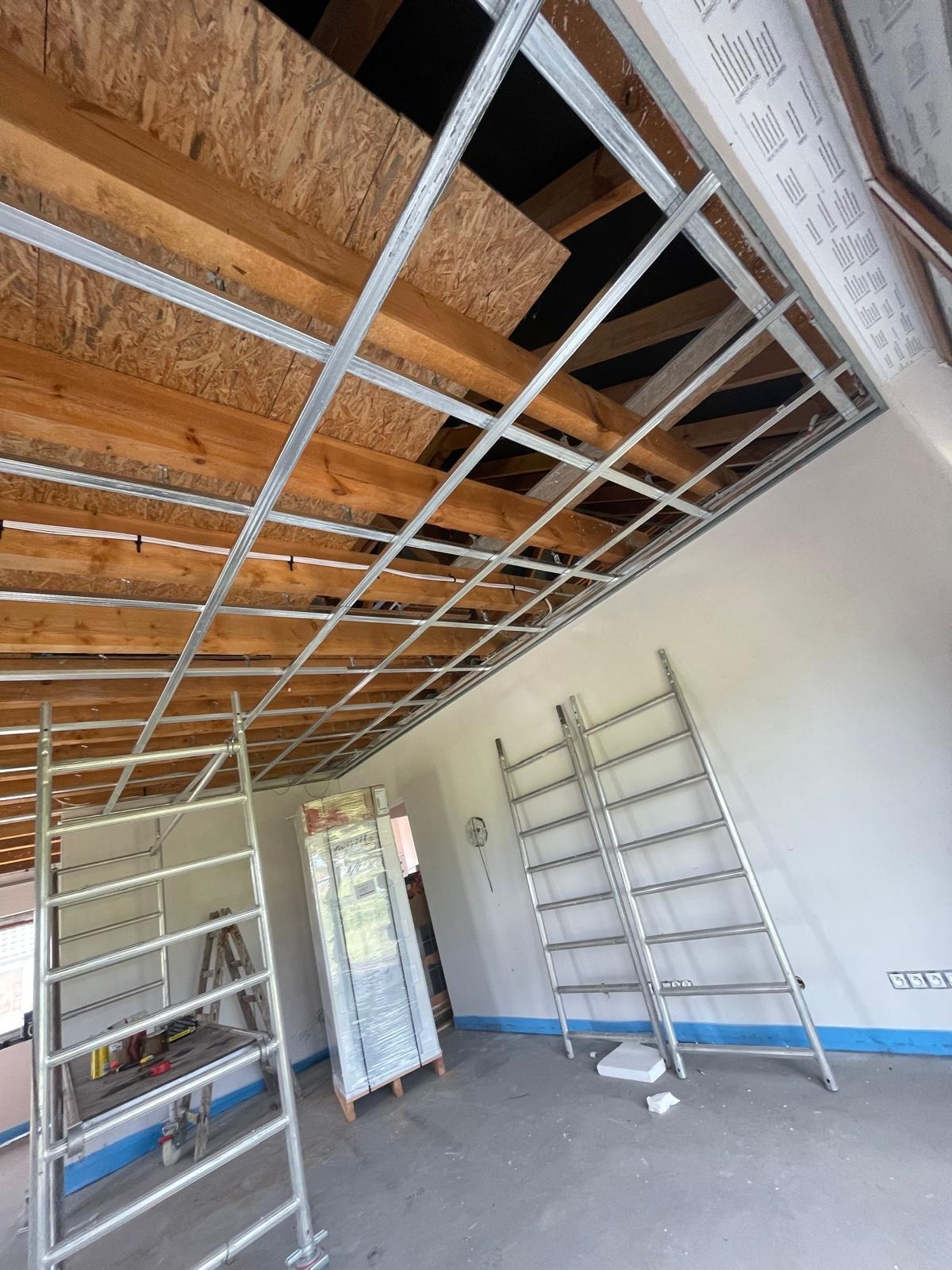 Montaż metalowego stelażu sufitu podwieszanego z widocznymi drewnianymi elementami konstrukcyjnymi, dwie rozstawione drabiny aluminiowe oraz zapakowane płyty karton-gips w folii.