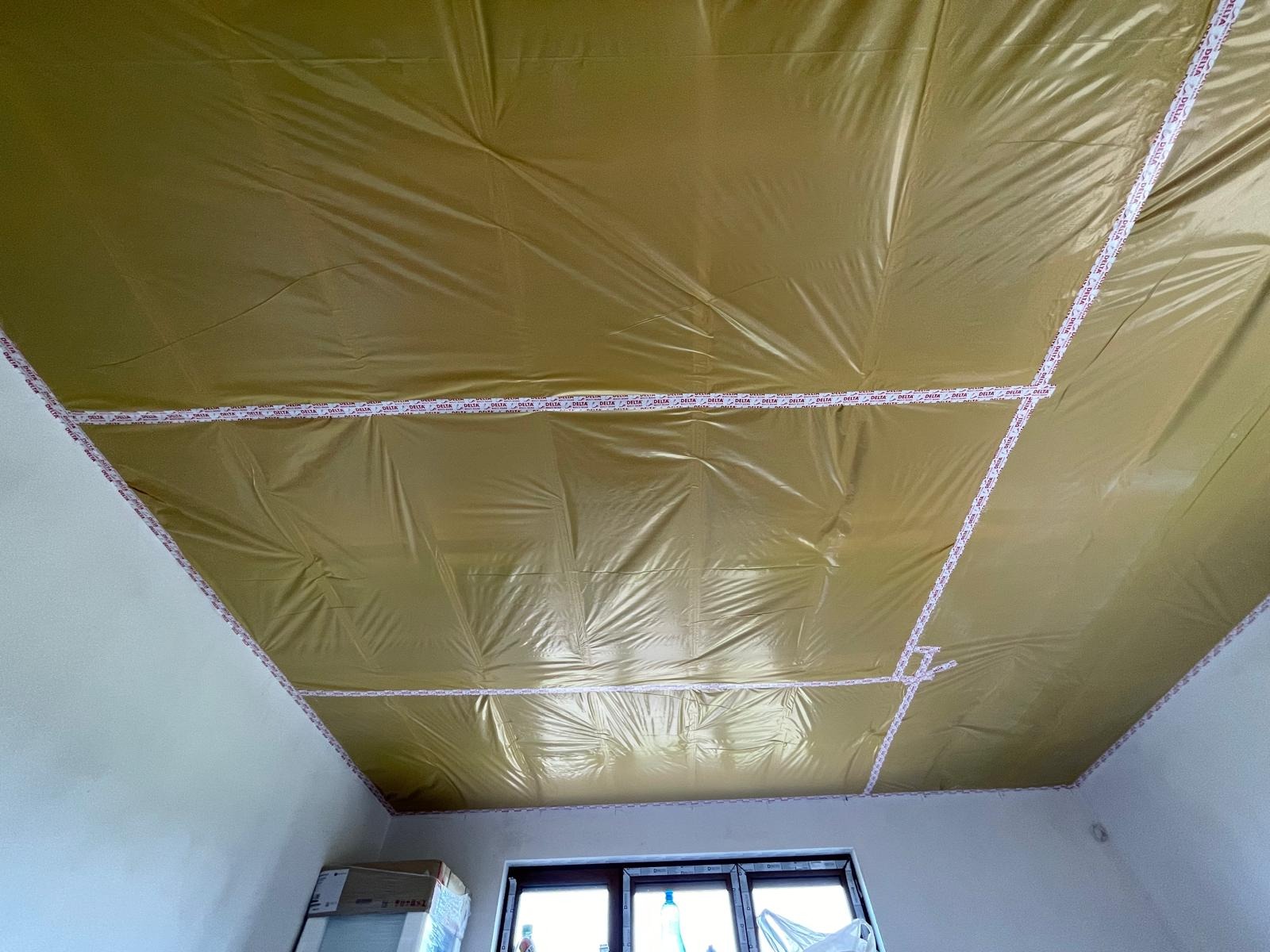 Folia paroizolacyjna na suficie poddasza, przyklejona taśmą Delta, widok z dołu na białe ściany i okno na tle nieba.