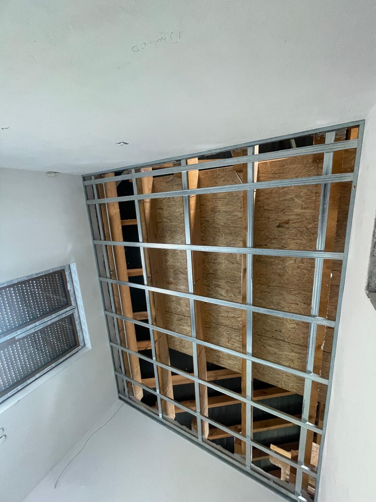 Metalowy stelaż pod płytę gipsowo-kartonową, widok od dołu, widoczne drewniane elementy konstrukcyjne oraz okno w tle.