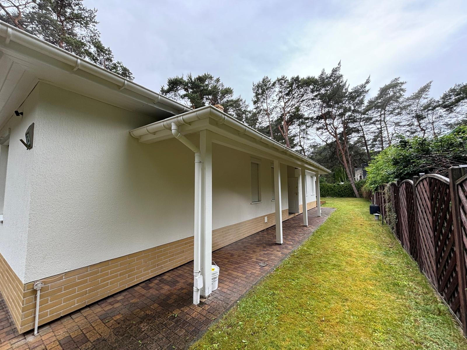 Jasny dom jednorodzinny z białymi kolumnami werandy i elewacją w odcieniach beżu, widok z boku na tle zielonego trawnika i drzew