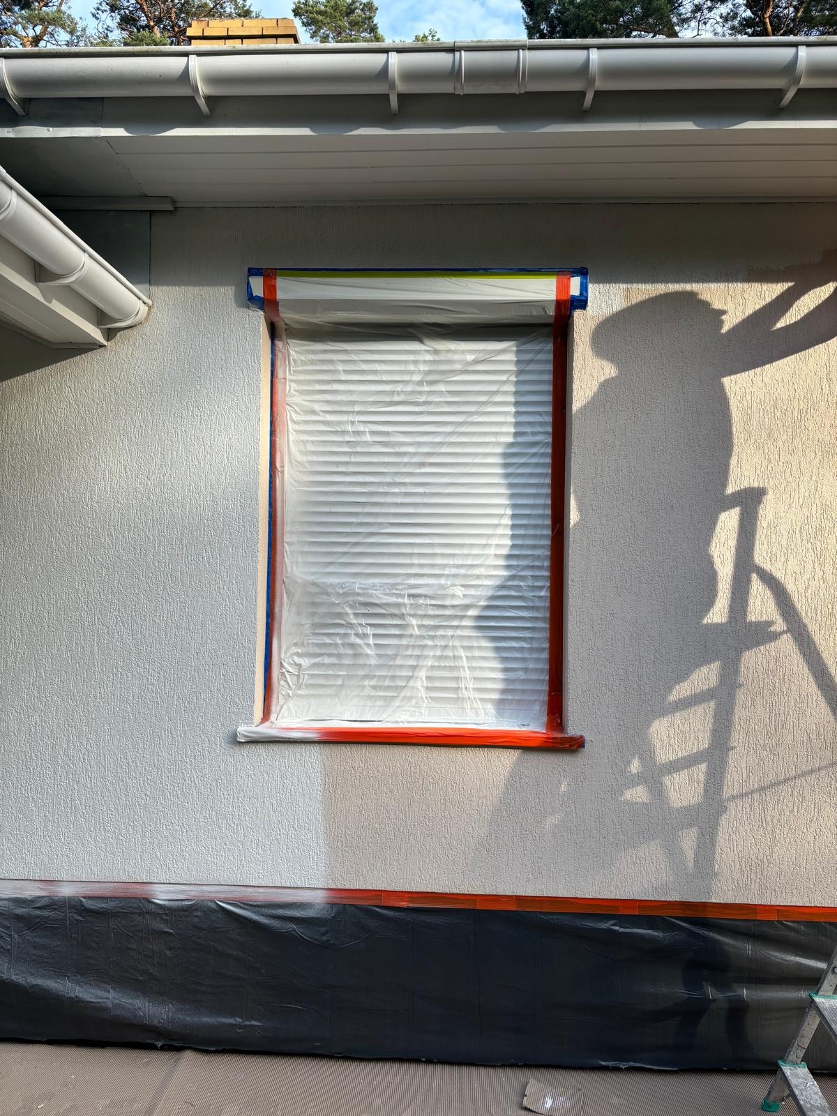 Przygotowanie elewacji do prac malarskich: okno zabezpieczone folią i taśmą, dolna część elewacji osłonięta czarną folią, widoczny cień osoby pracującej na drabinie.