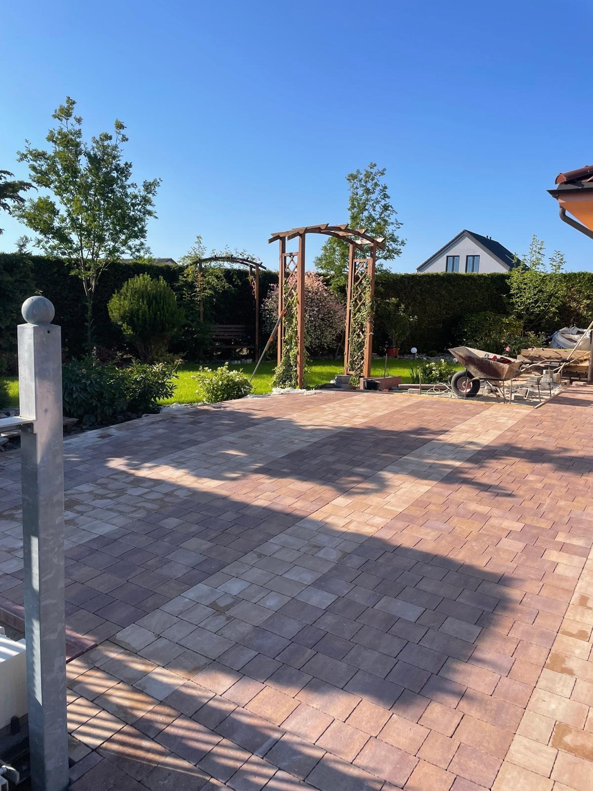 Ułożona kostka brukowa w ogrodzie z drewnianymi pergolami, w tle zielony żywopłot i dom.