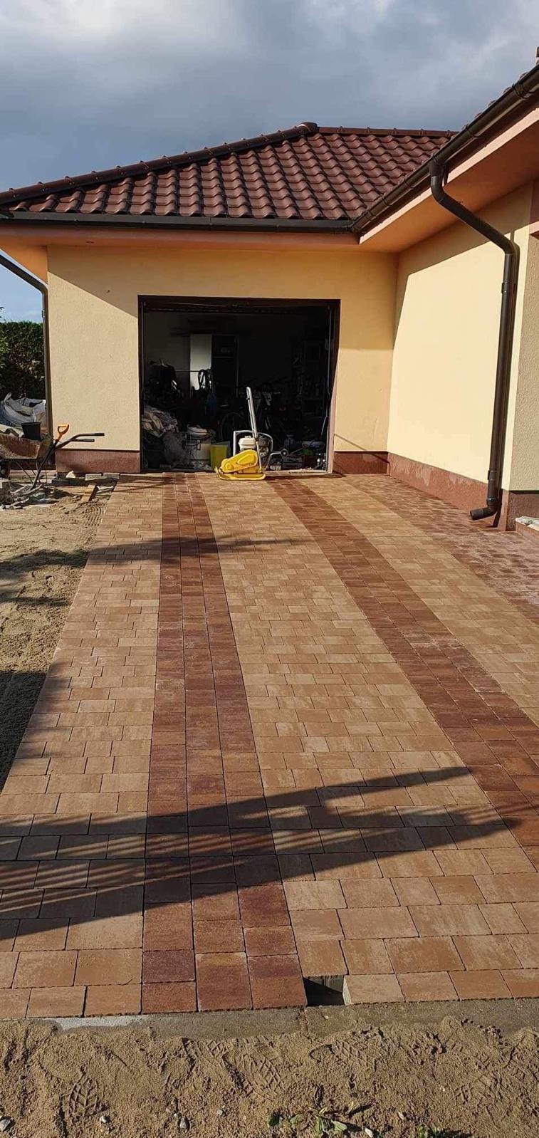 Ułożona kostka brukowa w dwóch odcieniach brązu tworząca wzór na podjeździe, widoczna zagęszczarka w otwartym garażu i piasek na obrzeżach.