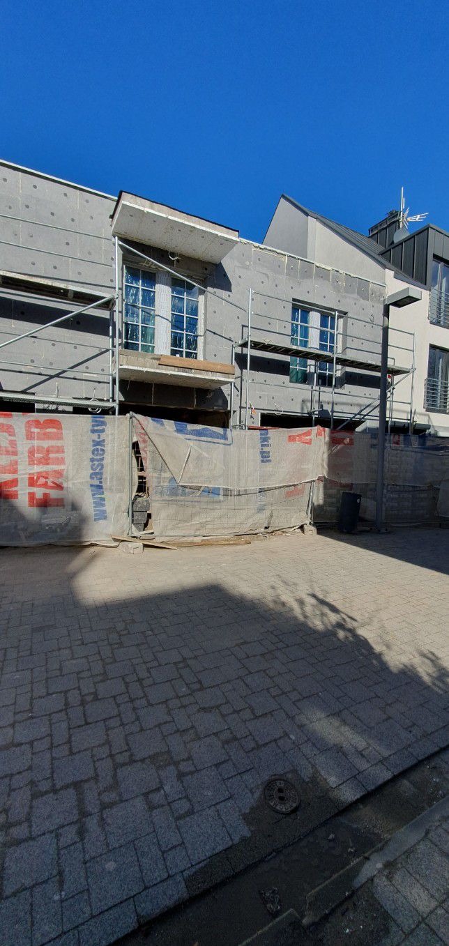 Budynek w trakcie prac elewacyjnych: ocieplenie ścian zewnętrznych widoczne na rusztowaniu, okna zabezpieczone folią, siatka osłonowa z reklamą na pierwszym planie.