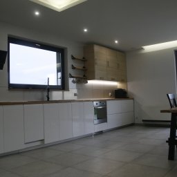 Dominik J - Nowoczesna kuchnia z białymi frontami szafek, drewnianym blatem i podświetlanym sufitem, widok na okno i część jadalni.