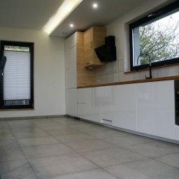 Dominik J - Nowoczesna kuchnia z białymi, lakierowanymi frontami szafek, drewnianymi akcentami i szarymi płytkami podłogowymi; minimalistyczny okap nad płytą grzewczą i czarna bateria przy oknie.