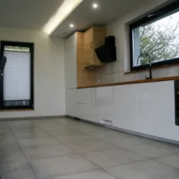 Nowoczesna kuchnia z białymi, lakierowanymi frontami szafek, drewnianymi akcentami i szarymi płytkami podłogowymi; minimalistyczny okap nad płytą grzewczą i czarna bateria przy oknie.