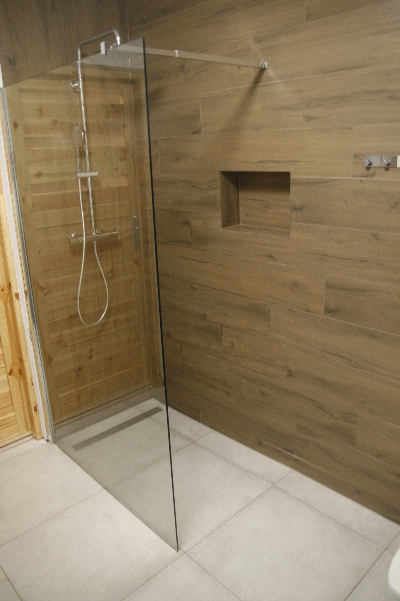 Kabina prysznicowa typu walk-in z deszczownicą, wykończona płytkami imitującymi drewno, z wnęką na kosmetyki w ścianie.