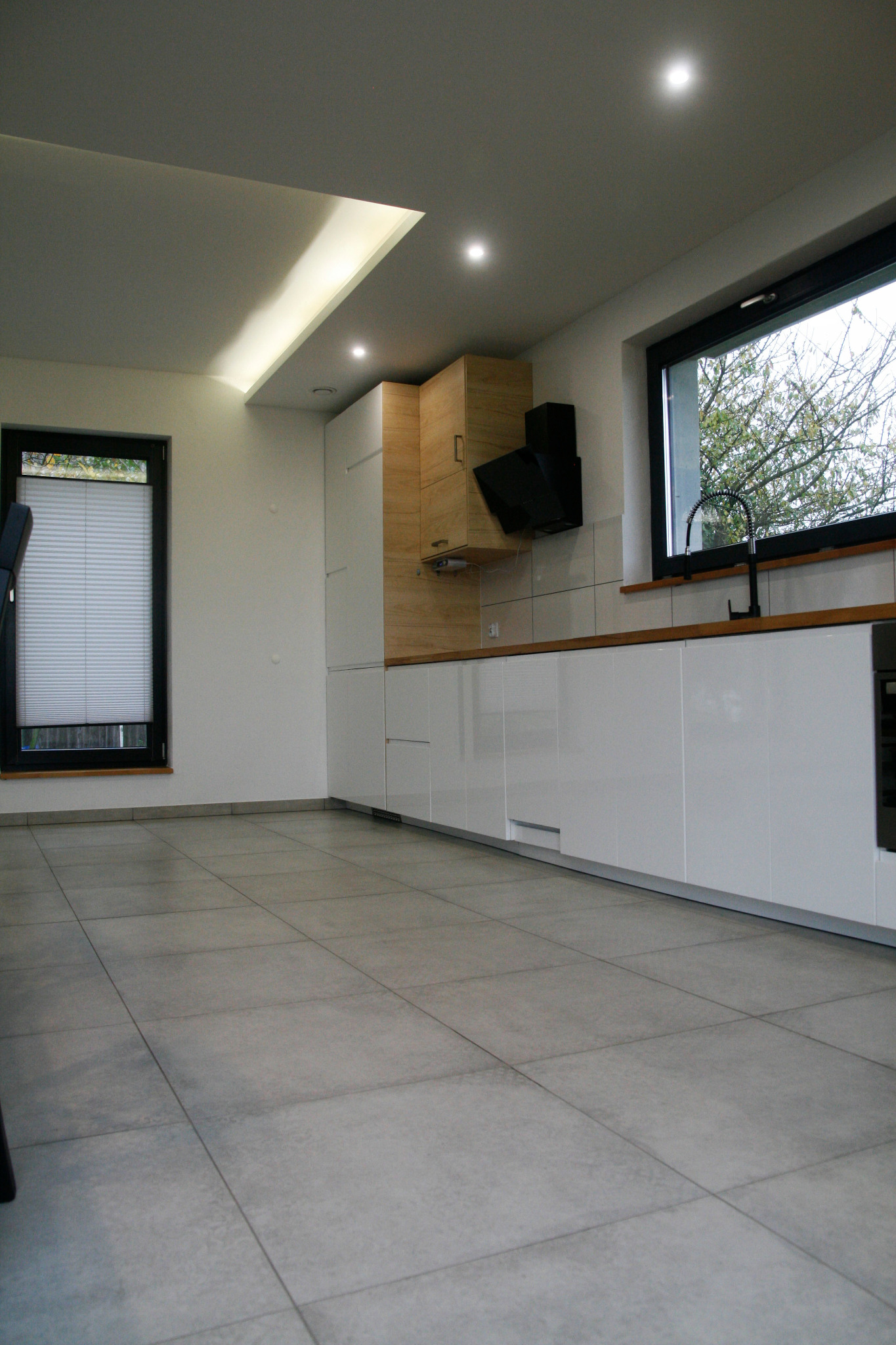 Nowoczesna kuchnia z białymi, lakierowanymi frontami szafek, drewnianymi akcentami i szarymi płytkami podłogowymi; minimalistyczny okap nad płytą grzewczą i czarna bateria przy oknie.