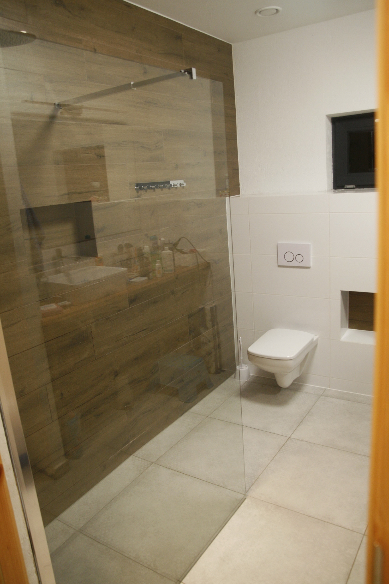 Nowoczesna łazienka z przeszkloną kabiną prysznicową i toaletą podwieszaną, wykończona płytkami imitującymi drewno i białymi płytkami, z widoczną armaturą i akcesoriami łazienkowymi.