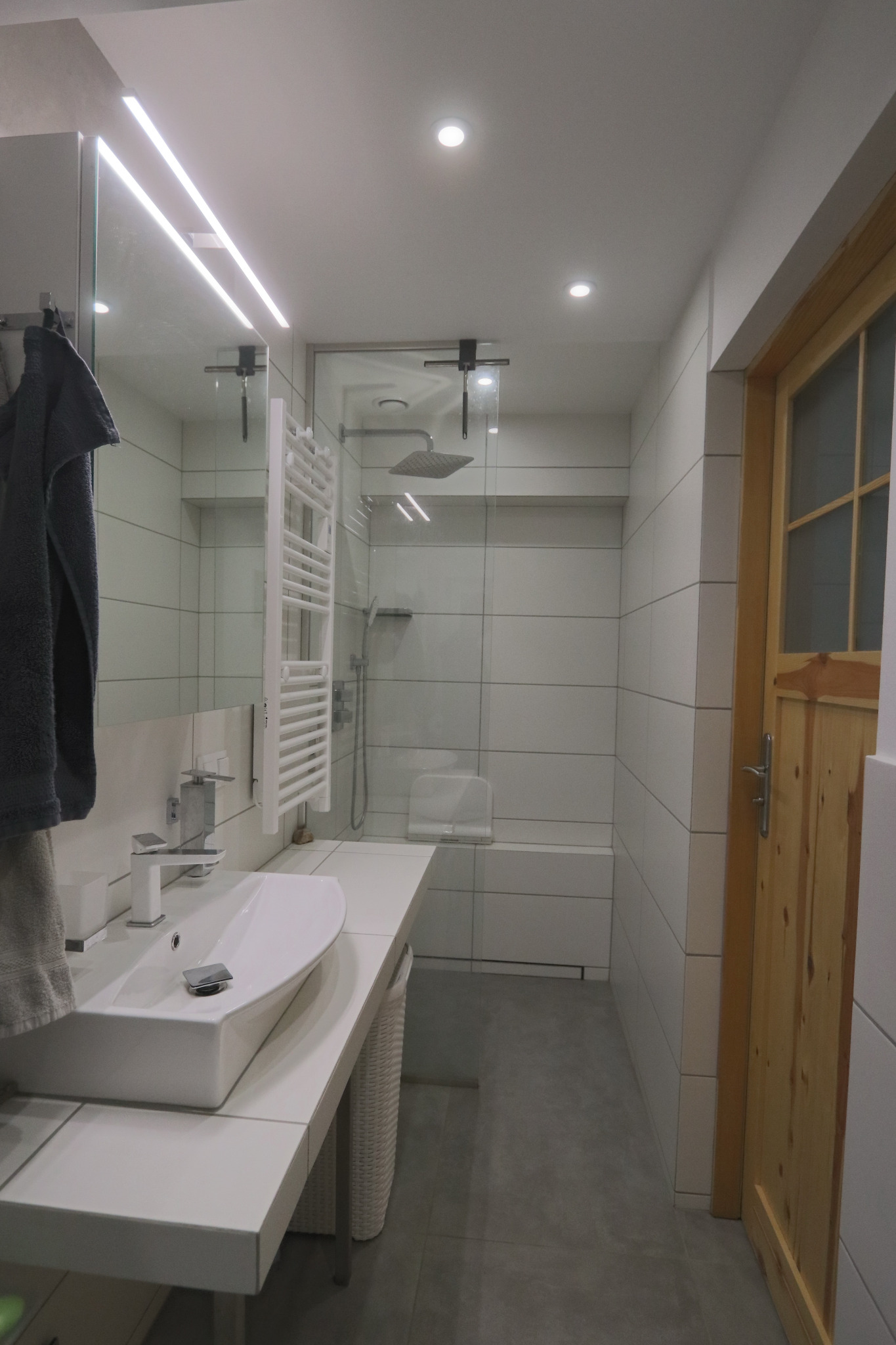 Nowoczesna łazienka z białą umywalką nablatową, grzejnikiem drabinkowym i kabiną prysznicową typu walk-in, oświetlona listwą LED w lustrze i drewnianymi drzwiami.