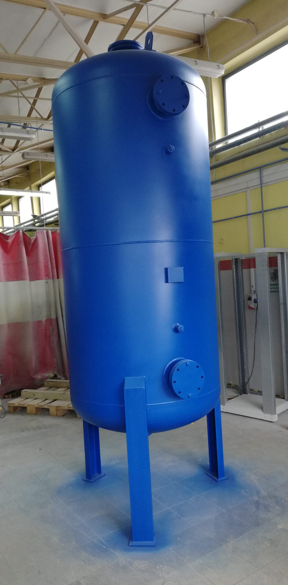 Wysoki, cylindryczny zbiornik ciśnieniowy w kolorze niebieskim, na trzech nogach, z widocznymi elementami przyłączeniowymi, świeżo pomalowany, z delikatną mgiełką farby na podłodze, w hali...