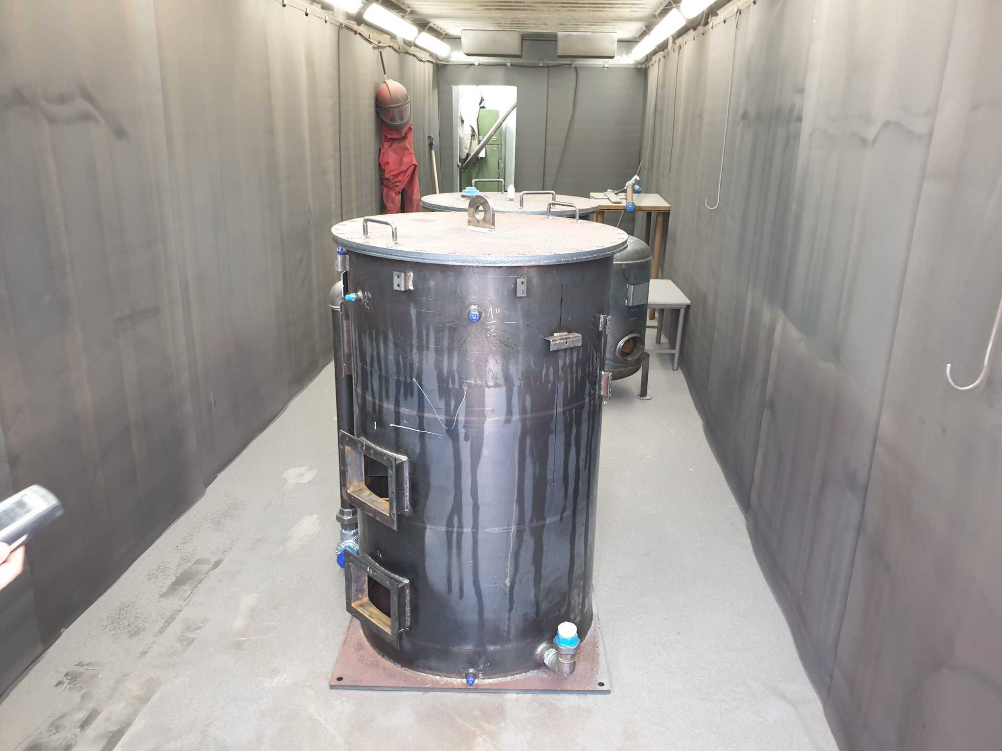 Metalowe zbiorniki wewnątrz pomieszczenia z szarymi zasłonami, widoczne ślady użytkowania i spawania, w tle sprzęt spawalniczy i osłona na ścianie.