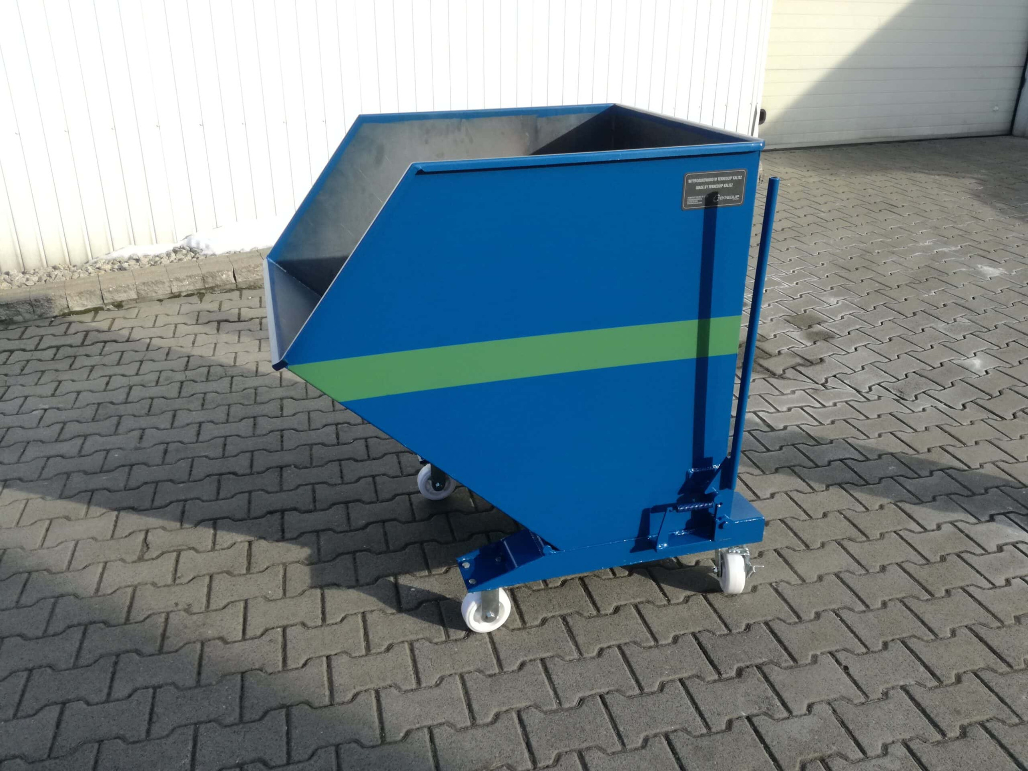 Niebieski kontener na kółkach z zielonym pasem, widoczny na tle kostki brukowej i białej ściany budynku.