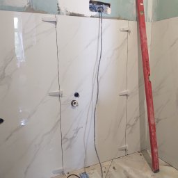 GregorBUD - Ściana w trakcie wykańczania z białymi płytkami imitującymi marmur, widoczne krzyżyki dystansowe, poziomica PRO oparta o ścianę i otwory na gniazdka elektryczne.