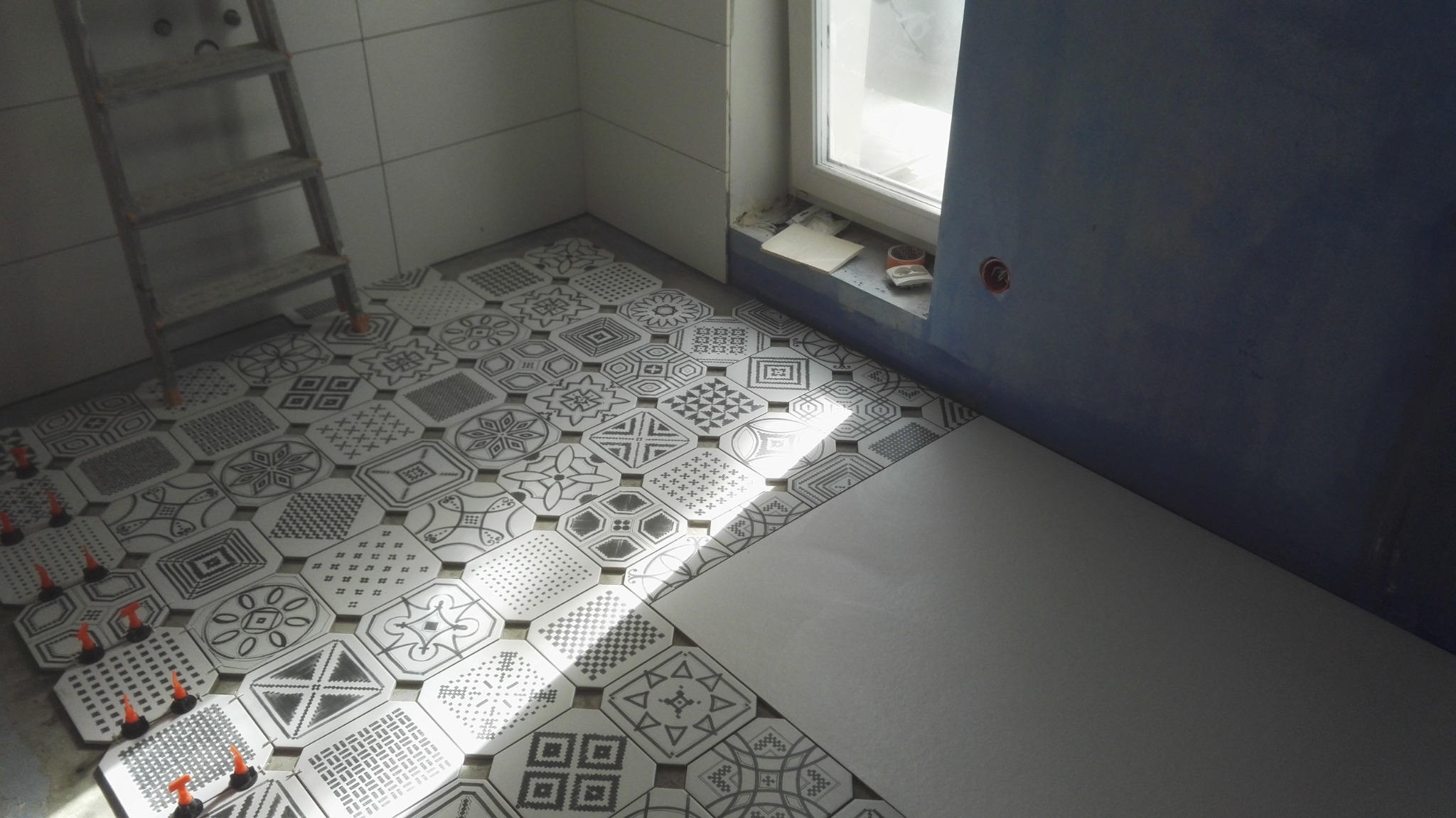 Wnętrze łazienki w trakcie układania wzorzystych, czarno-białych płytek podłogowych; widoczna drabina, okno, oraz fragment ściany przygotowanej pod malowanie