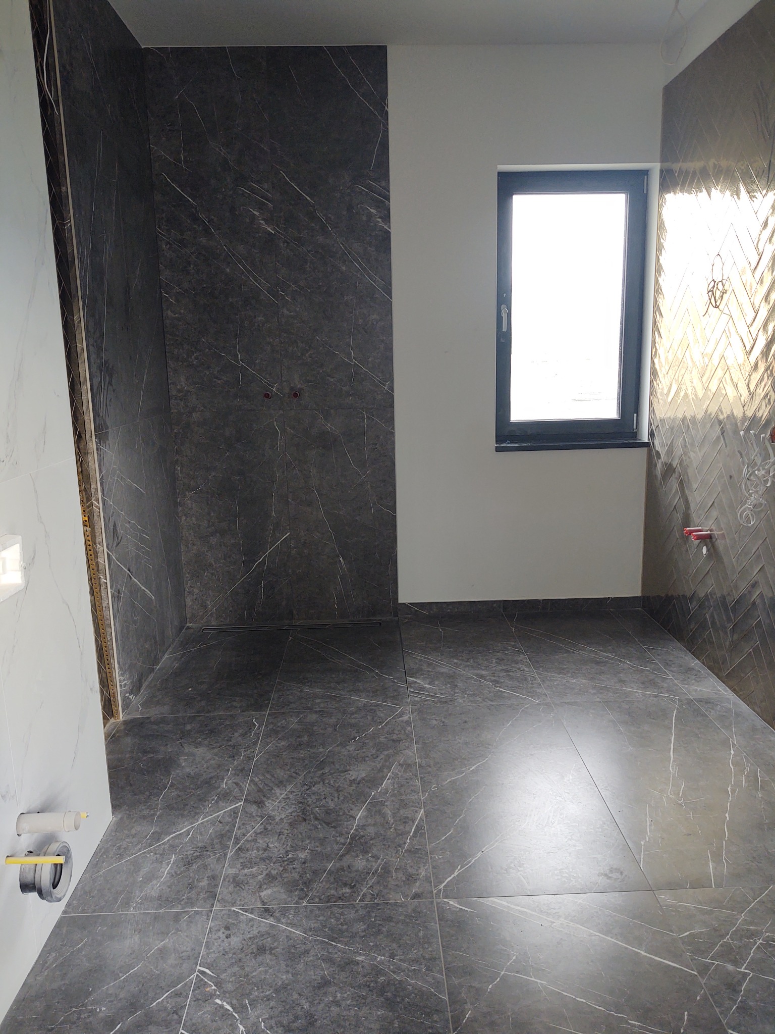 Łazienka w trakcie wykańczania: szare płytki imitujące marmur na podłodze i ścianach, jedna ściana wykończona w jodełkę, widoczne punkty instalacji wodnej i okno.