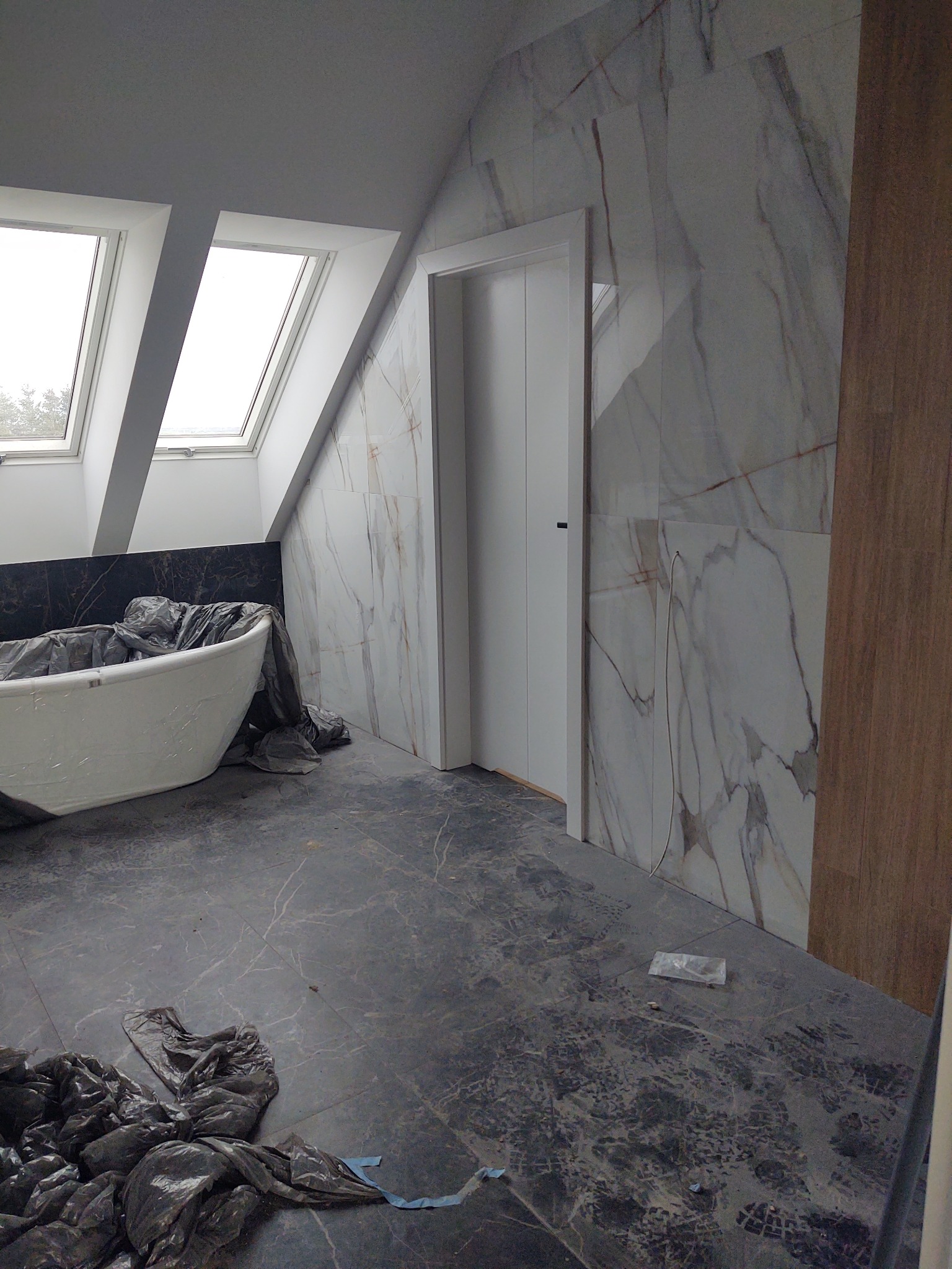 Łazienka na poddaszu w trakcie wykańczania: marmurowe płytki na ścianach, zabezpieczona folią wanna, widoczne okna dachowe i ślady po pracach na podłodze.