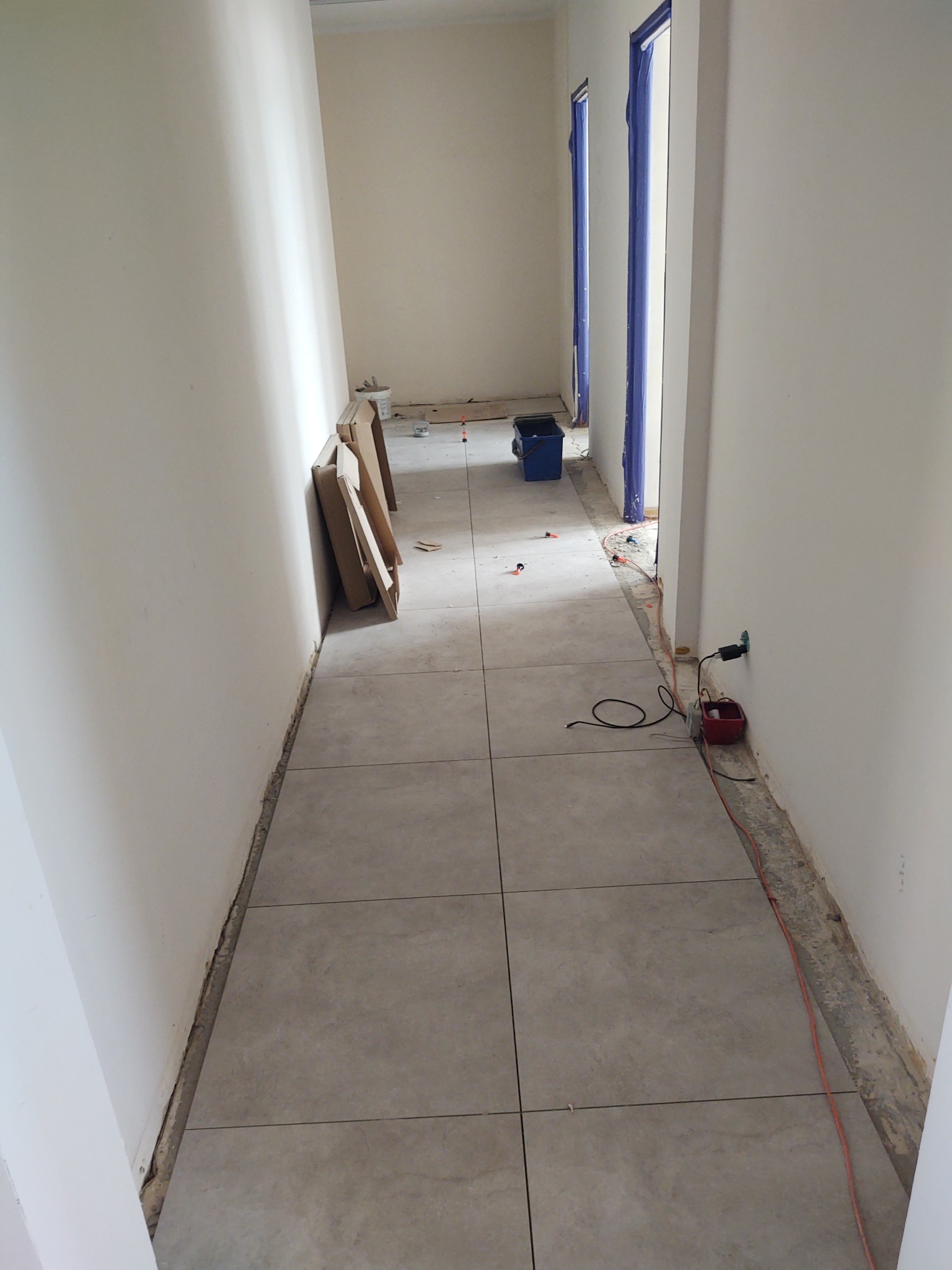 Korytarz w trakcie układania szarych płytek podłogowych, widoczne narzędzia, zabezpieczone folią drzwi i fragment nie wykończonej ściany przy podłodze.