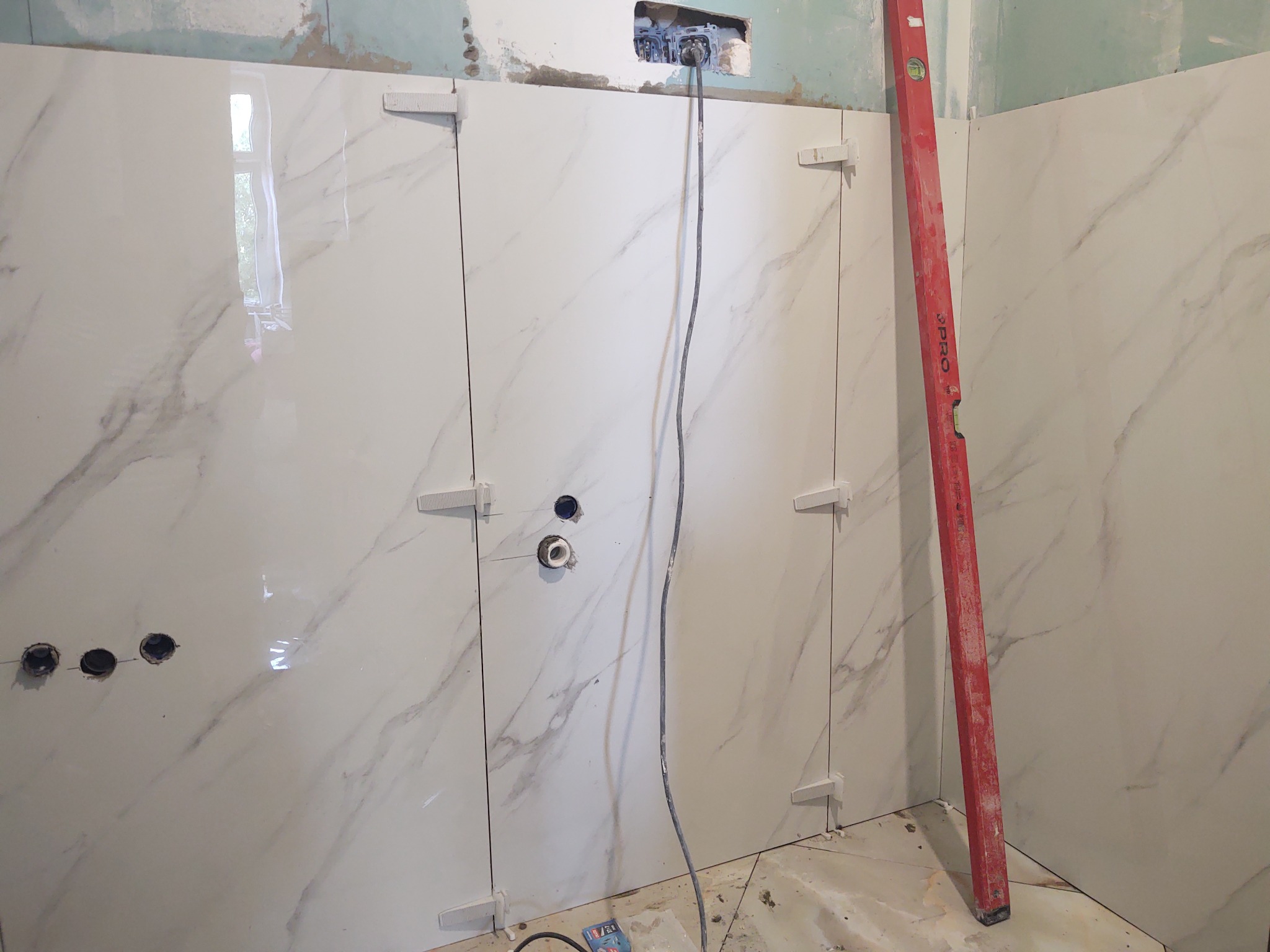 Ściana w trakcie wykańczania z białymi płytkami imitującymi marmur, widoczne krzyżyki dystansowe, poziomica PRO oparta o ścianę i otwory na gniazdka elektryczne.