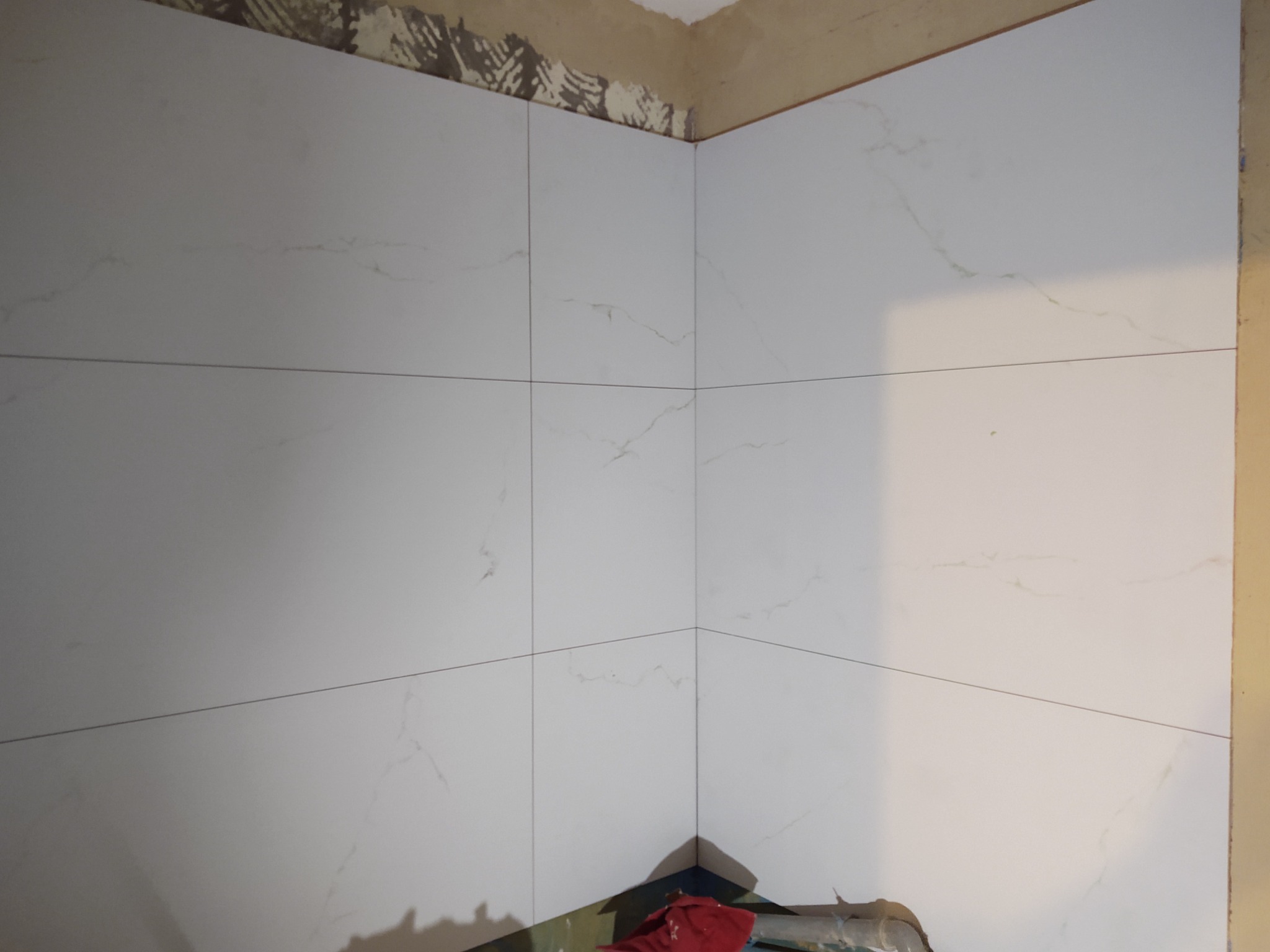 Fragment ściany w rogu pomieszczenia z ułożonymi białymi płytkami imitującymi marmur, widoczne niedociągnięcia na górze ściany i narzędzia w dolnym rogu.