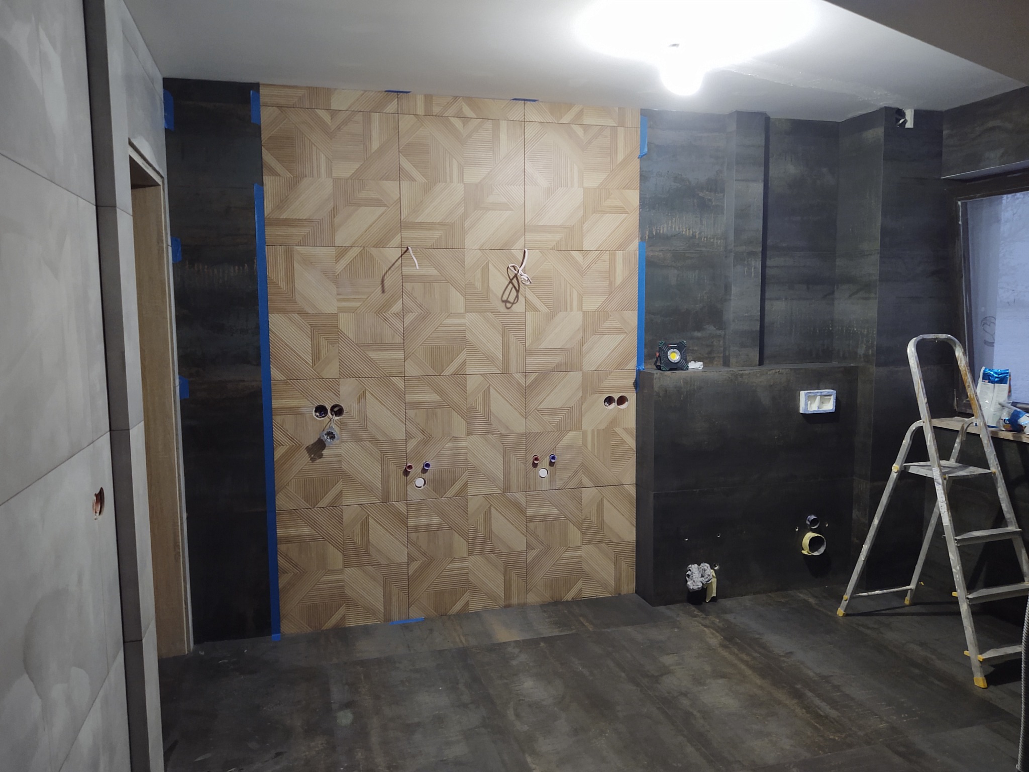 Ściana w trakcie wykańczania z dekoracyjnymi panelami drewnopodobnymi w geometryczny wzór, widoczne niepodłączone kable i otwory na gniazdka, zabezpieczenia taśmą malarską na styku z ciemnoszarą...