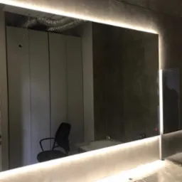 Prostokątne lustro z podświetleniem LED w łazience z szarymi płytkami, odbijające szafy, krzesło i instalację wentylacyjną pod sufitem.