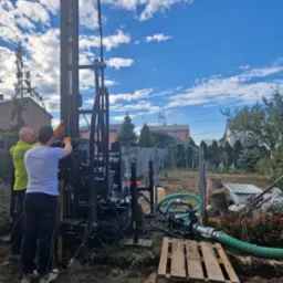 Wiercenie studni głębinowej w Ładzicach: dwaj mężczyźni obsługują wiertnicę na tle błękitnego nieba z obłokami, widoczna pompa wodna i teren budowy.