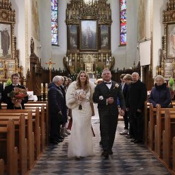 Bogusława Trela Fotografia - Para młoda w kościele, panna młoda w sukni z cekinami i futrzanym okryciem, pan młody w kraciastej marynarce, goście weselni w tle, drewniane ławki kościelne.