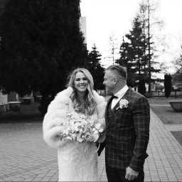 Bogusława Trela Fotografia - Czarno-białe zdjęcie uśmiechniętej panny młodej w futrze i sukni ślubnej z bukietem, stojącej obok pana młodego w garniturze w kratę, na tle brukowanej ścieżki i drzew