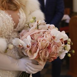 Bogusława Trela Fotografia - Panna młoda w futrzanym szalu trzyma bukiet z różowych róż i białych orchidei, w tle fragment wnętrza kościoła.
