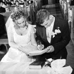 Bogusława Trela Fotografia - Czarno-białe ujęcie pary młodej podpisującej akt małżeństwa w kościele, panna młoda w koronkowej sukni z perłami, pan młody w ciemnym garniturze z butonierką, w tle widoczni goście.