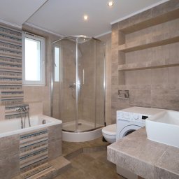 Bogusława Trela Fotografia - Jasna łazienka z wanną, kabiną prysznicową, umywalką nablatową i pralką, wykończona beżowymi płytkami z poziomymi paskami.