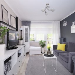 Bogusława Trela Fotografia - Jasne, nowocześnie urządzone wnętrze salonu z szarą sofą z żółtą poduszką, białymi meblami, roślinami doniczkowymi i szarym dywanem.