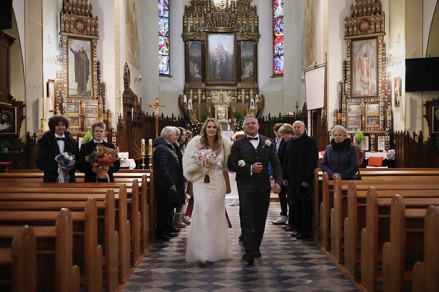 Para młoda w kościele, panna młoda w sukni z cekinami i futrzanym okryciem, pan młody w kraciastej marynarce, goście weselni w tle, drewniane ławki kościelne.