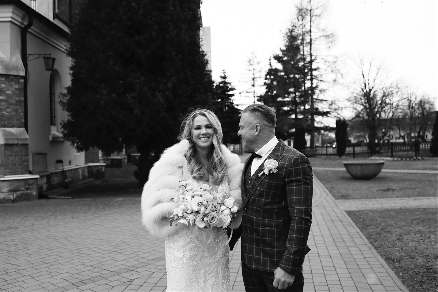 Czarno-białe zdjęcie uśmiechniętej panny młodej w futrze i sukni ślubnej z bukietem, stojącej obok pana młodego w garniturze w kratę, na tle brukowanej ścieżki i drzew