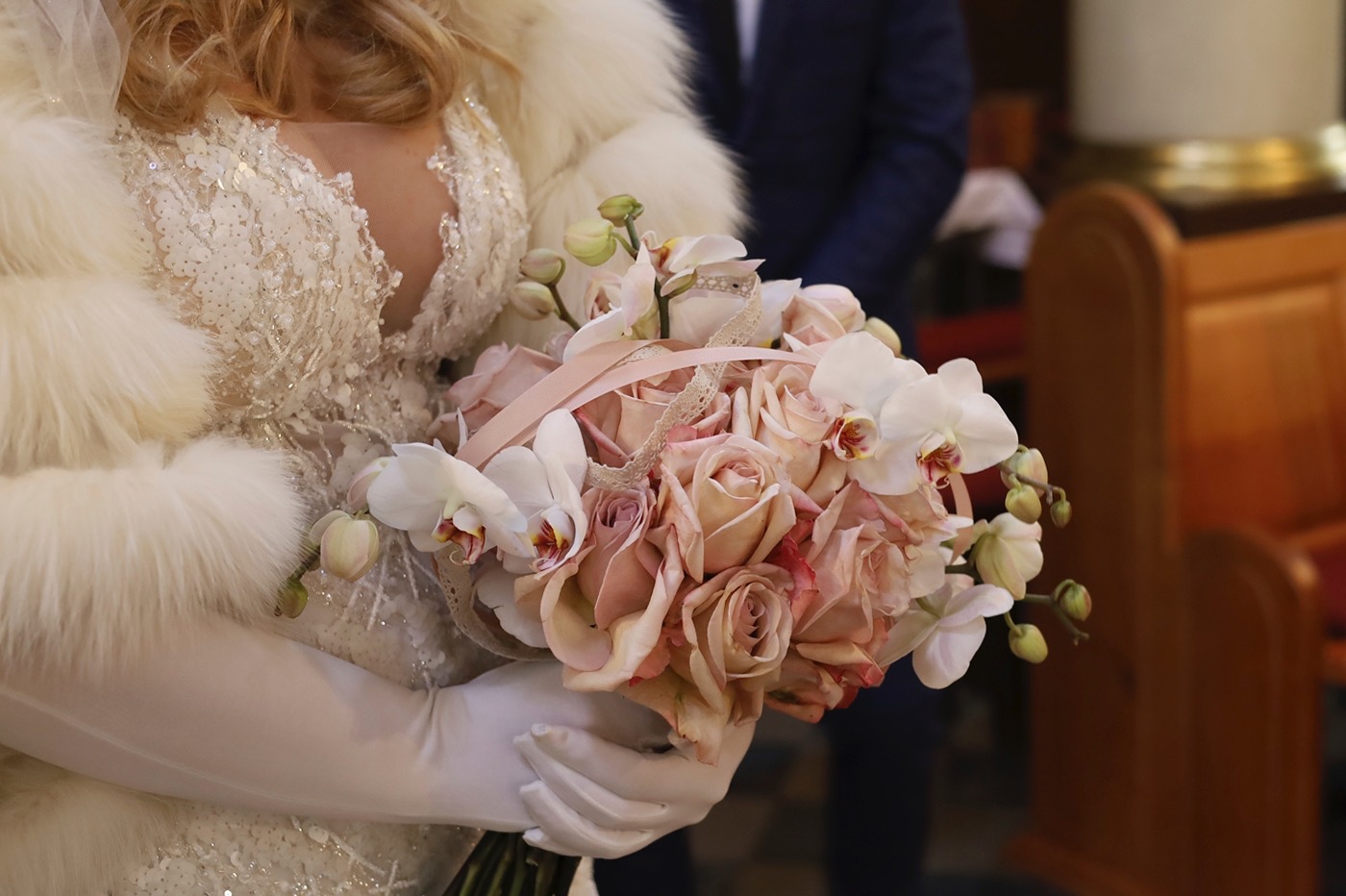 Panna młoda w futrzanym szalu trzyma bukiet z różowych róż i białych orchidei, w tle fragment wnętrza kościoła.