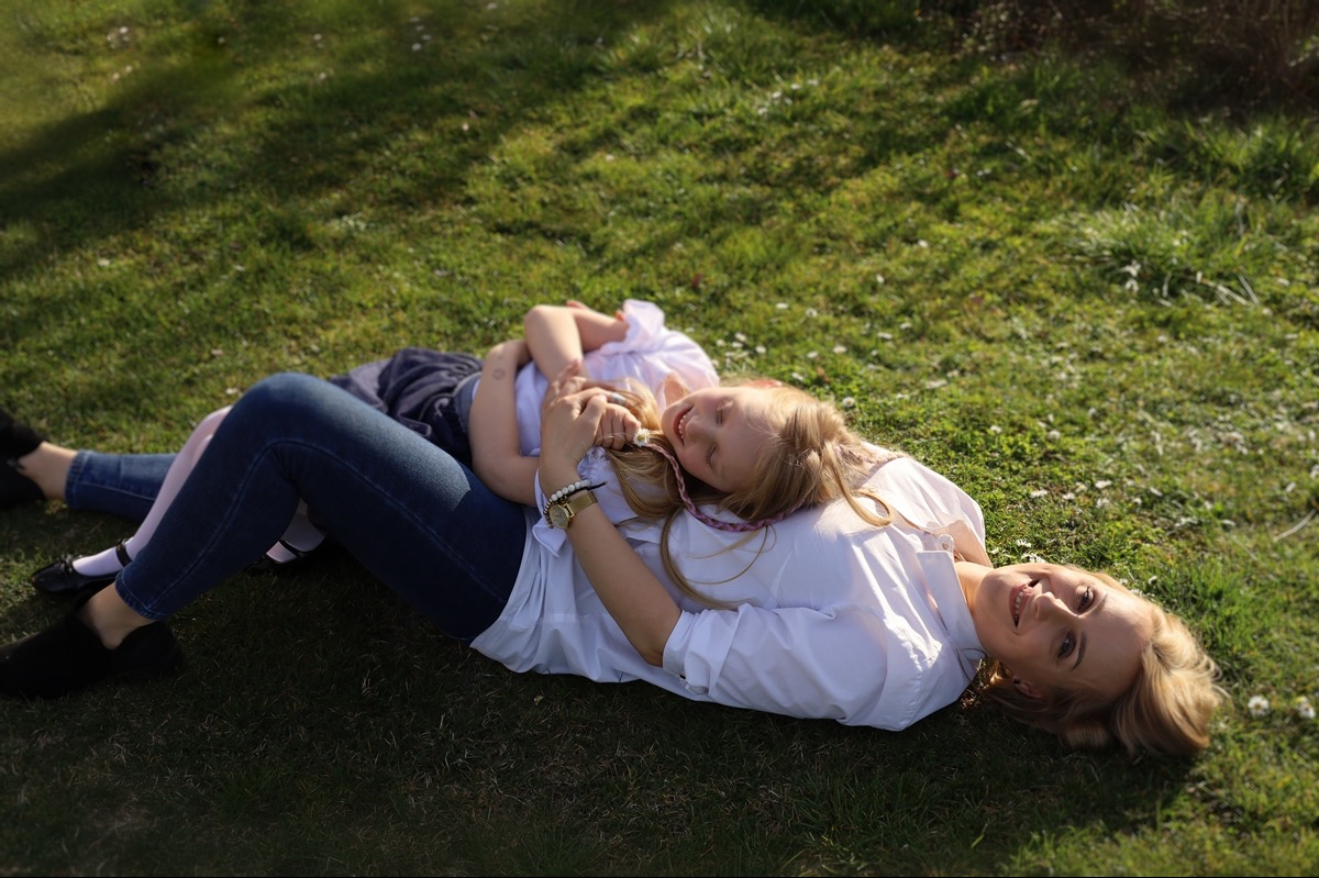 Mama i córka leżą na trawie, córka opiera głowę na mamie, obie uśmiechnięte, w tle zielona trawa i cień drzewa.