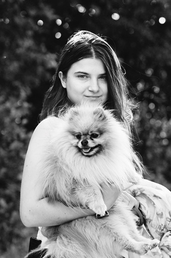 Portret kobiety z długimi włosami trzymającej na rękach małego psa rasy szpic miniaturowy, wykonany w czerni i bieli na tle rozmytej zieleni.